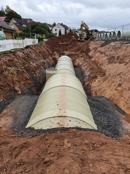 Bauarbeiten an einer großen Rohrleitung auf einer Baustelle, mit einem Bagger im Hintergrund und weiter entfernten Häusern.