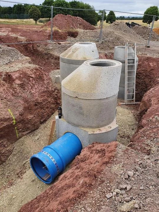 Bauarbeiten an einer Klärgrube mit Betonrohren, Leiter und Baufahrzeugen auf einer Baustelle im Freien.