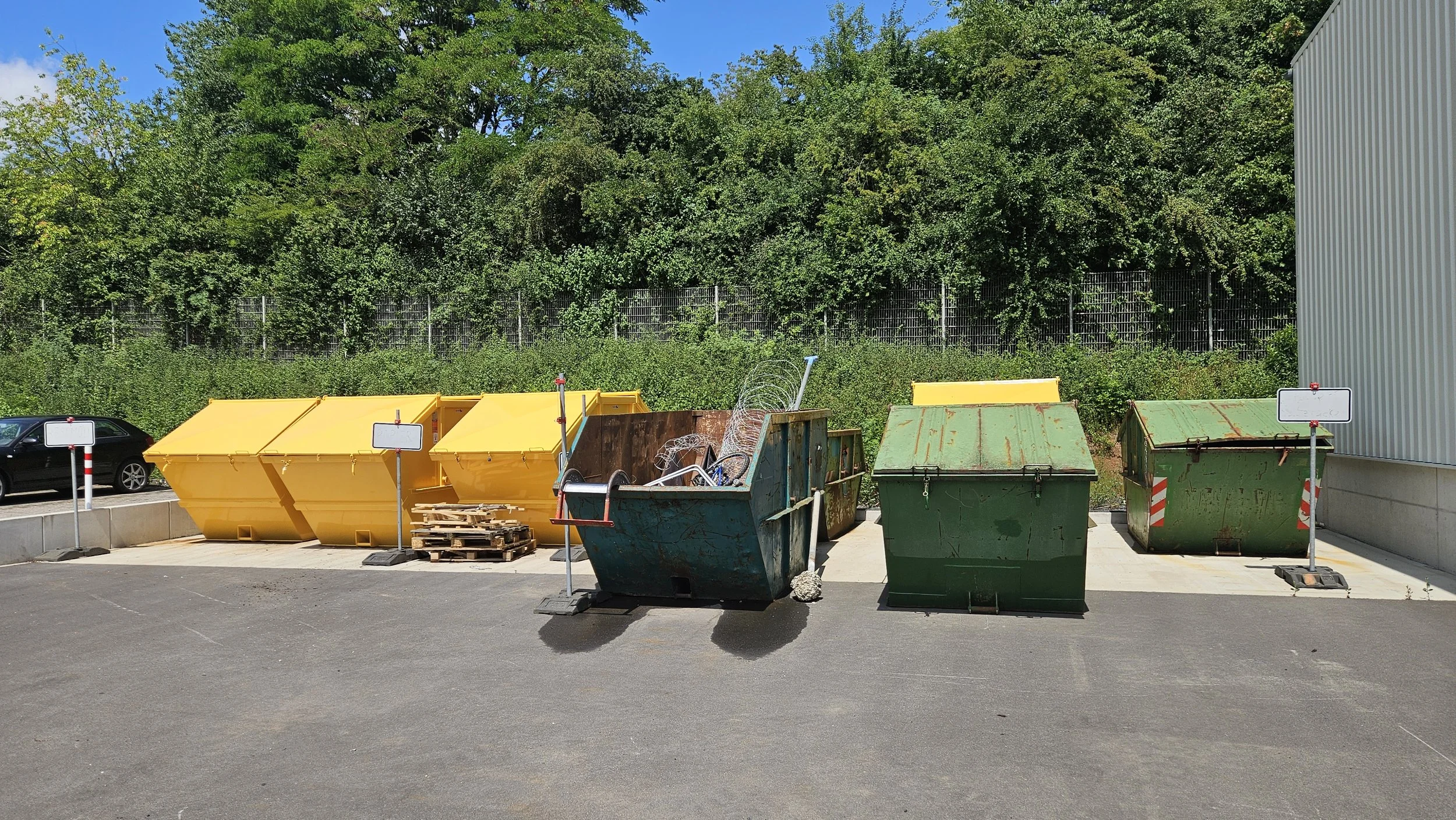 Fünf große Abfalldosen, größtenteils grün und gelb, stehen in einer Parklücke vor einem Zaun und einem Gebäude, umgeben von einem asphaltierten Parkplatz, mit Bäumen im Hintergrund.