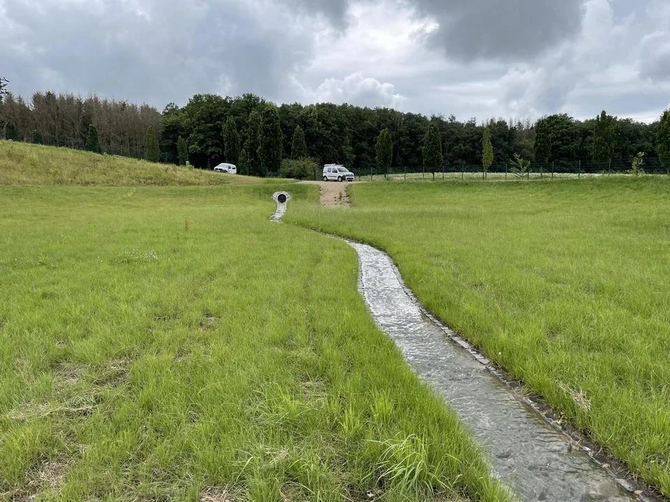 Ein kleines Wasserbächlein schlängelt sich durch eine grüne Wiesenlandschaft, im Hintergrund sind Bäume und zwei weiße Fahrzeuge. Die Wolken am Himmel sind bewölkt.