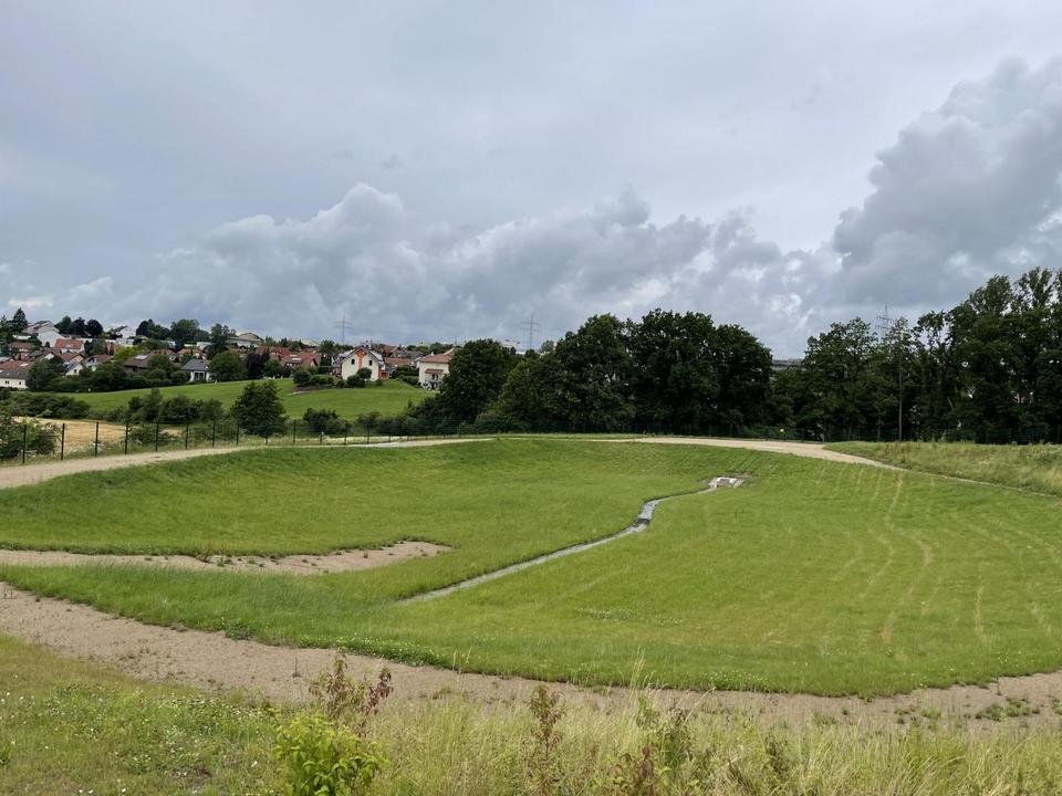 Grünes, ebenes Feld mit einem kleinen Wasserlauf, im Hintergrund ein Wohngebiet mit Häusern und Bäumen, bewölkter Himmel.