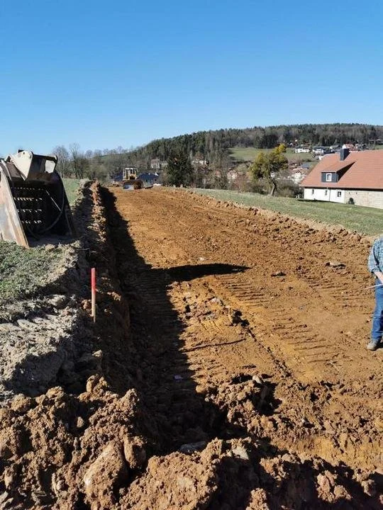 Ein landwirtschaftliches Bauprojekt mit Erdarbeiten, Traktor im Hintergrund, eine Person mit Messgerät auf der rechten Seite, unter klarem blauen Himmel.