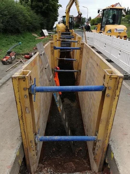 Baugrube mit seitlichen Stützwänden und beweglichen Schraubverbänden, im Hintergrund Baumaschinen und Bauarbeiter auf einer Straße.