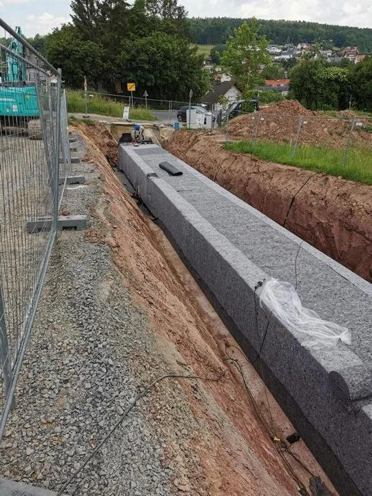 Bauarbeiten an einer Straße, mit einem langen, aufgeständerten Betonelement in einem Graben, umgeben von Erde, Steinen und einem Bauzaun.