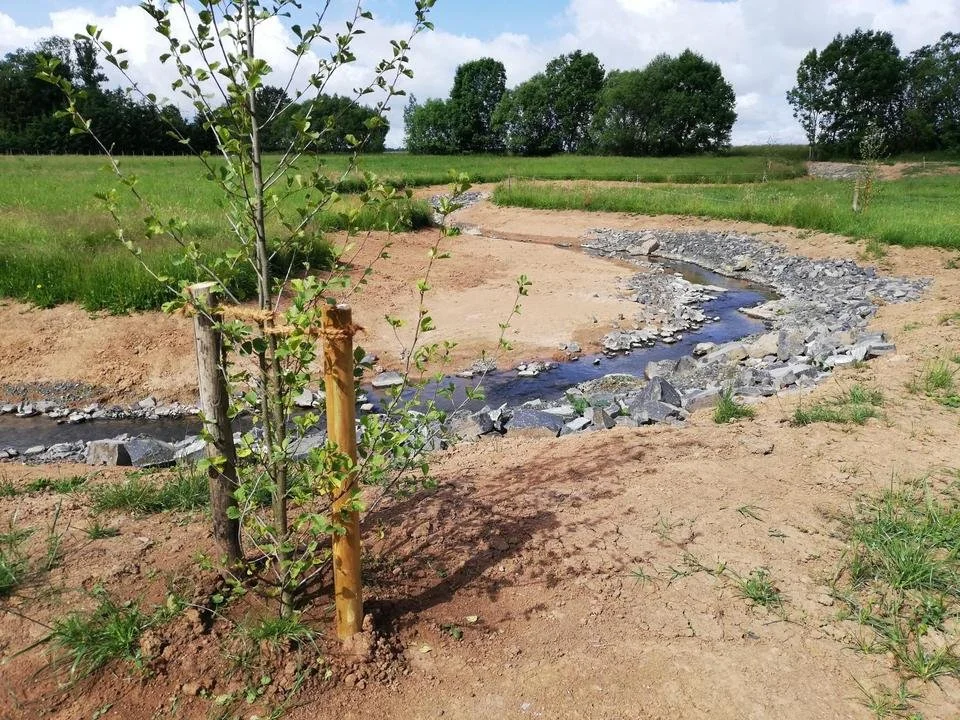Kleiner Bach in einer landwirtschaftlichen Umgebung, umgeben von Erde und Gras, mit einigen Bäumen im Hintergrund und einem wolkigen Himmel.