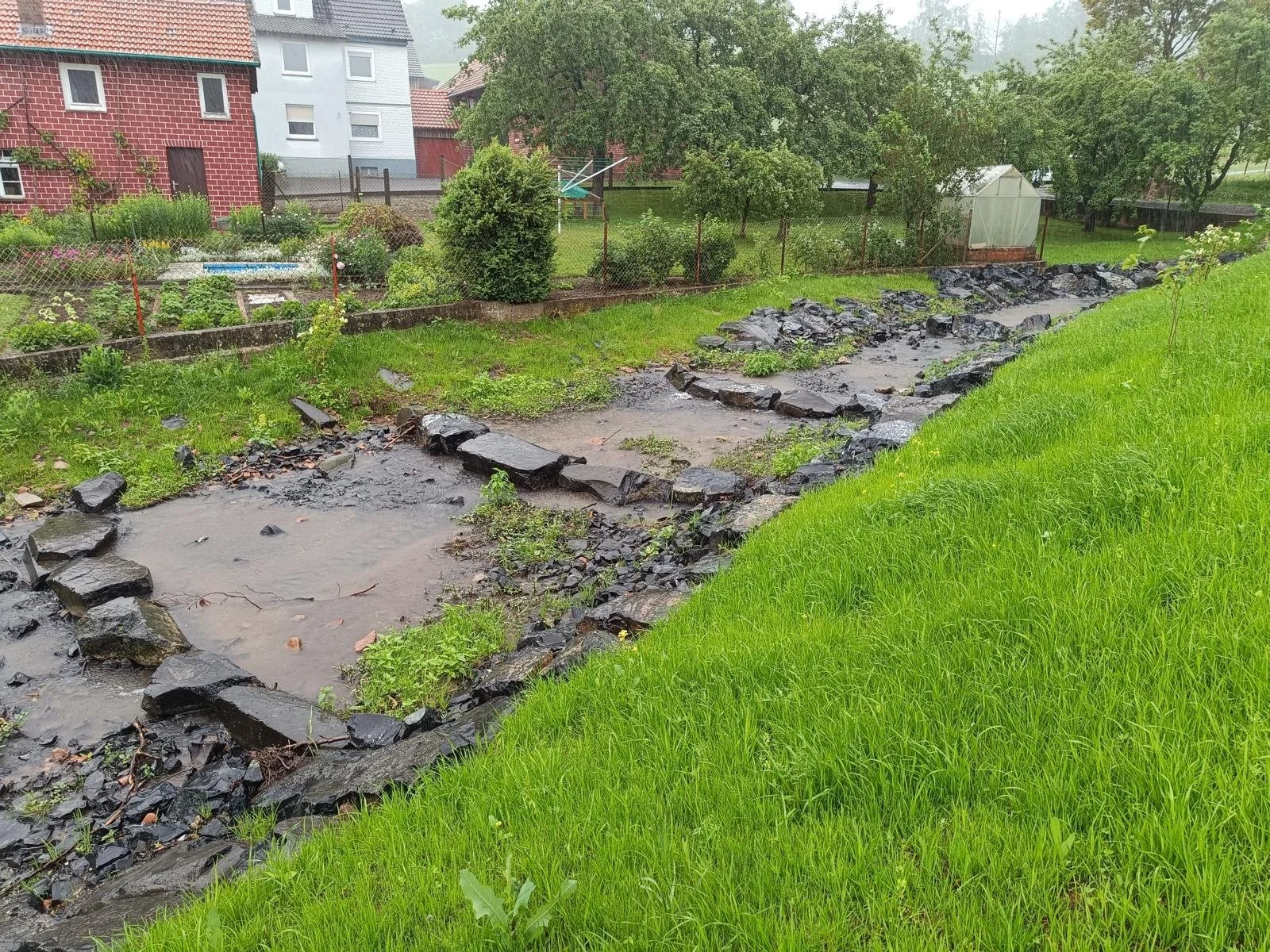 Ein grasbewachsener Garten mit einem kleinen Wasserlauf und Steinen auf einer Seite, mit Bäumen, Häusern und einem Gartengebäude im Hintergrund bei Regen.