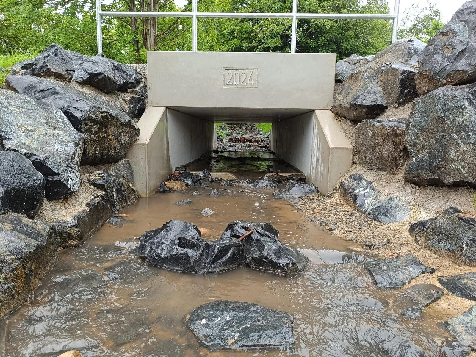 Ein Wasserkanal mit Steinen, umgeben von Felsen und Bäumen, mit der Jahreszahl 2024 auf einer Betonbrücke darüber.