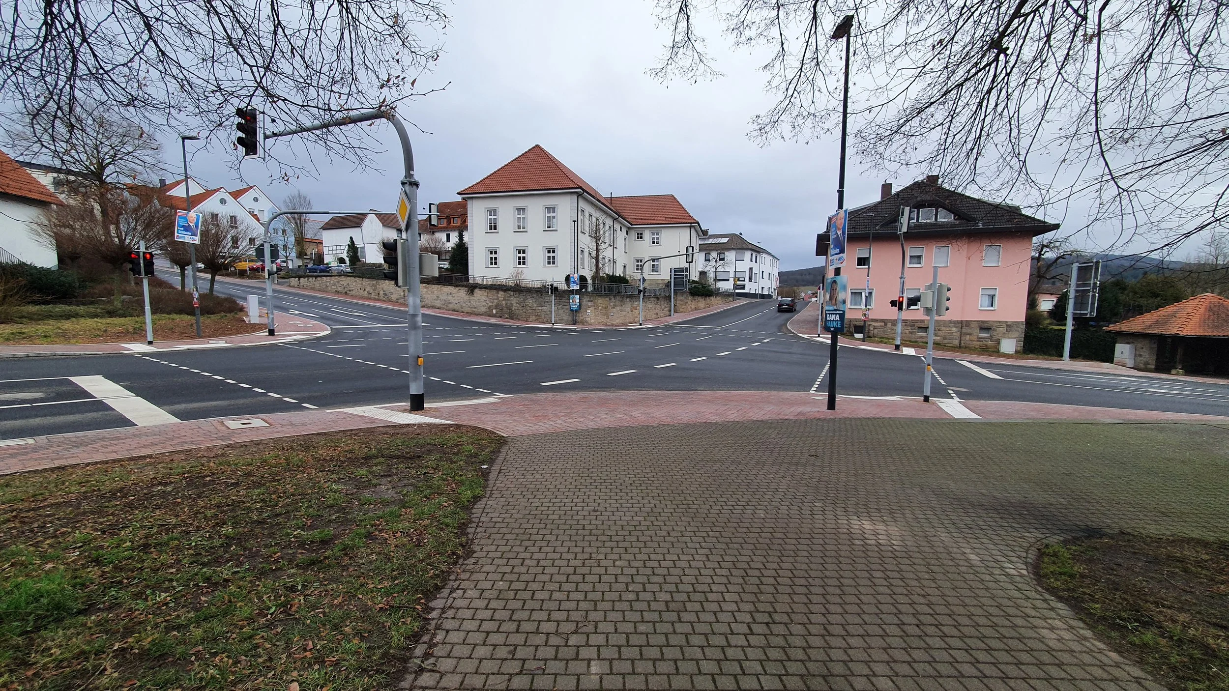 Blick auf eine mehrspurige Kreuzung in einer Wohngegend mit mehreren weißen und rosa Häusern, Ampeln und Verkehrsschildern bei bewölktem Himmel.