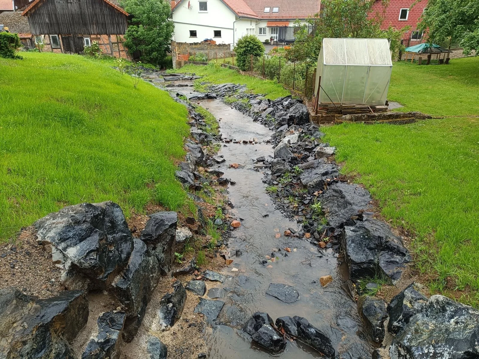 Ein kleiner Bach, umgeben von grünen Rasenflächen und Steinen, mit Häusern im Hintergrund, eines davon eine Gartenhütte, bei Regenwetter.