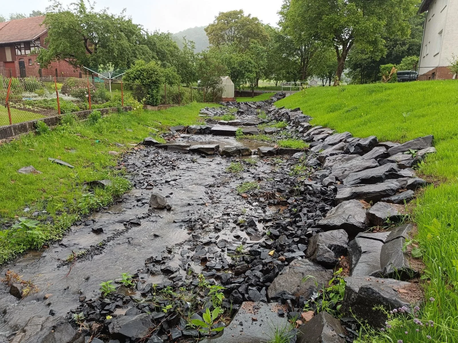 Ein kleiner Bach fließt durch ein grünes, bewachsenes Gelände, umgeben von Steinen und Gras. Im Hintergrund sind Häuser, Bäume und ein bewölkter Himmel zu sehen.