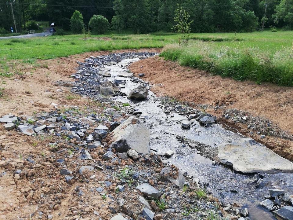 Ein kleiner Wasserlauf oder Bach in einem offenen Bereich, gesäumt von Erde und Kies, mit grünen Wiesen und Bäumen im Hintergrund.