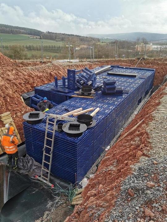 Baustelle mit blauen Kunststoffrohren im Erdgrab, Bauarbeiter tragen orange Jacken, ländliche Umgebung im Hintergrund