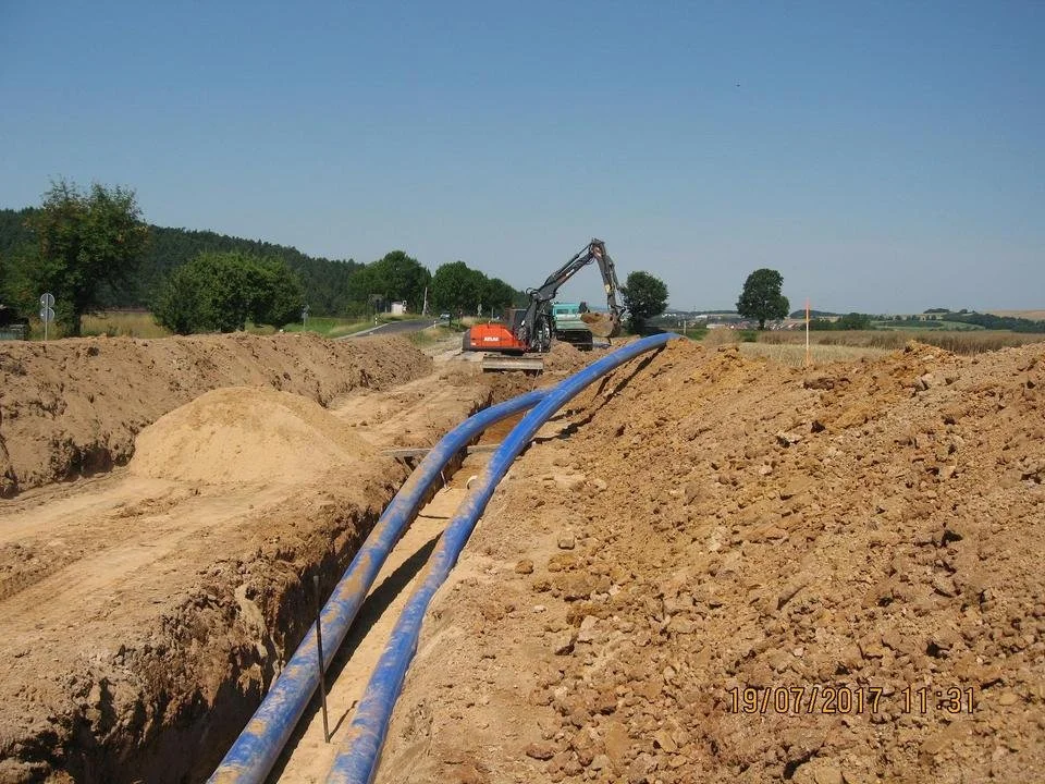 Bauarbeiter verlegt blaue Wasserleitungen in einen Graben auf einer Baustelle in einer ländlichen Gegend bei sonnigem Wetter.