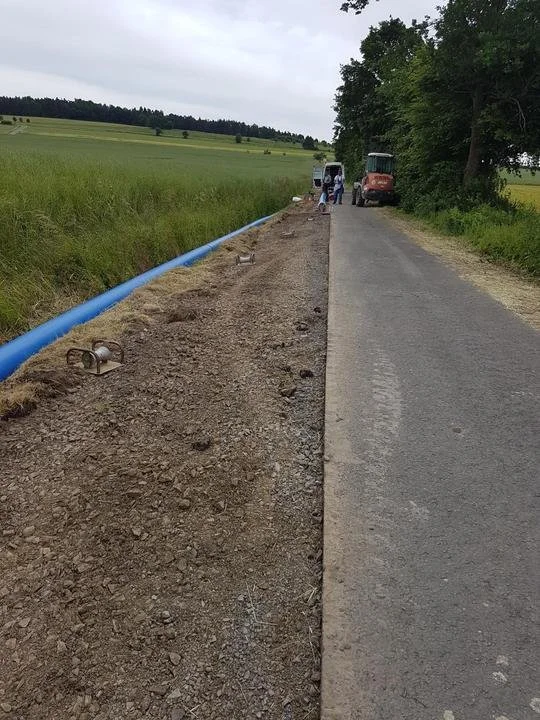 Ein landwirtschaftlicher Weg mit einer blauen Rohrleitung auf der linken Seite und Baustellenarbeiten auf der rechten Seite, mit einem Baumaschinen und Arbeitern im Hintergrund.