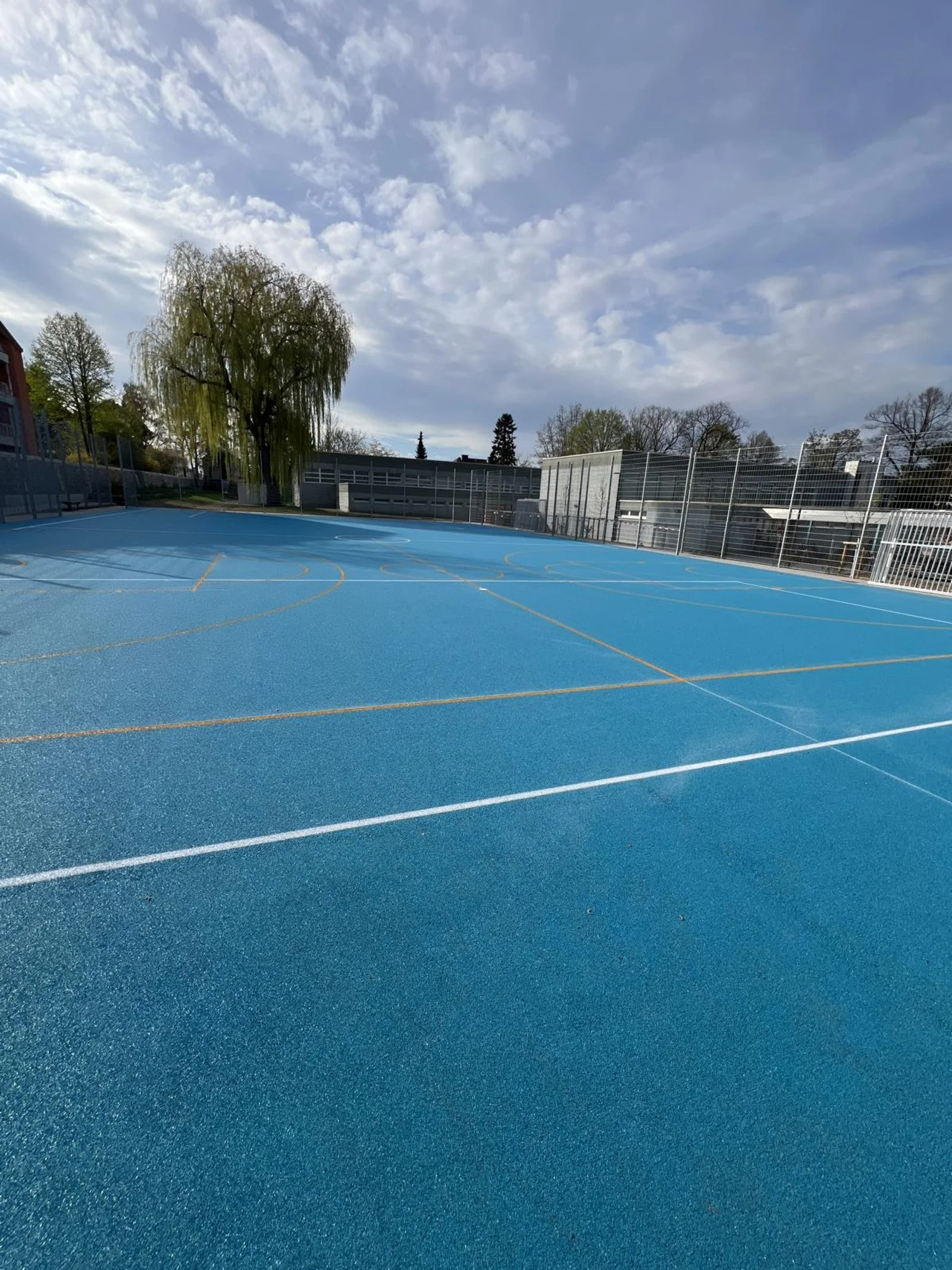 Leerer blauer Tennisplatz im Freien mit weißen Linien, umgeben von einem Zaun, bei sonnigem Himmel.