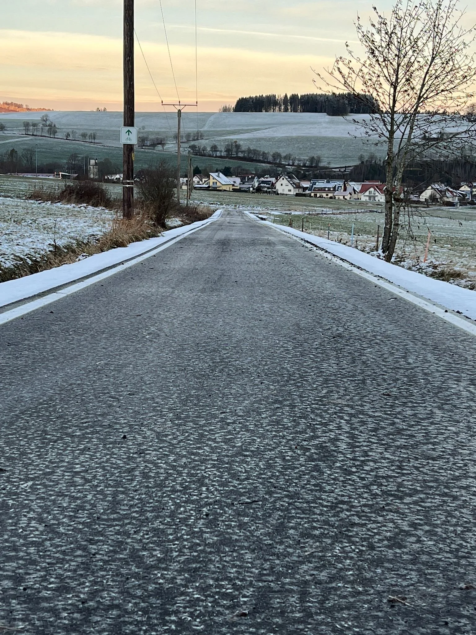 Eine ländliche Straße im Winter mit Schnee an den Seiten, Häuser im Hintergrund und eine Baum auf der rechten Seite.