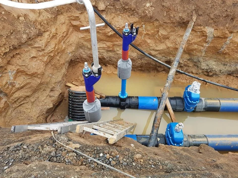 Bauarbeiten an einer Wasserleitung im Boden, mit blauen und schwarzen Rohren, Wasser im Graben, und Werkzeugen