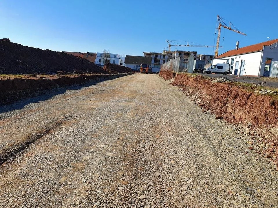 Straßenbaustelle mit unbefestigtem, schottrigem Weg, Bauarbeiter und schwere Baumaschinen, im Hintergrund sind Gebäude und Baukräne sichtbar.