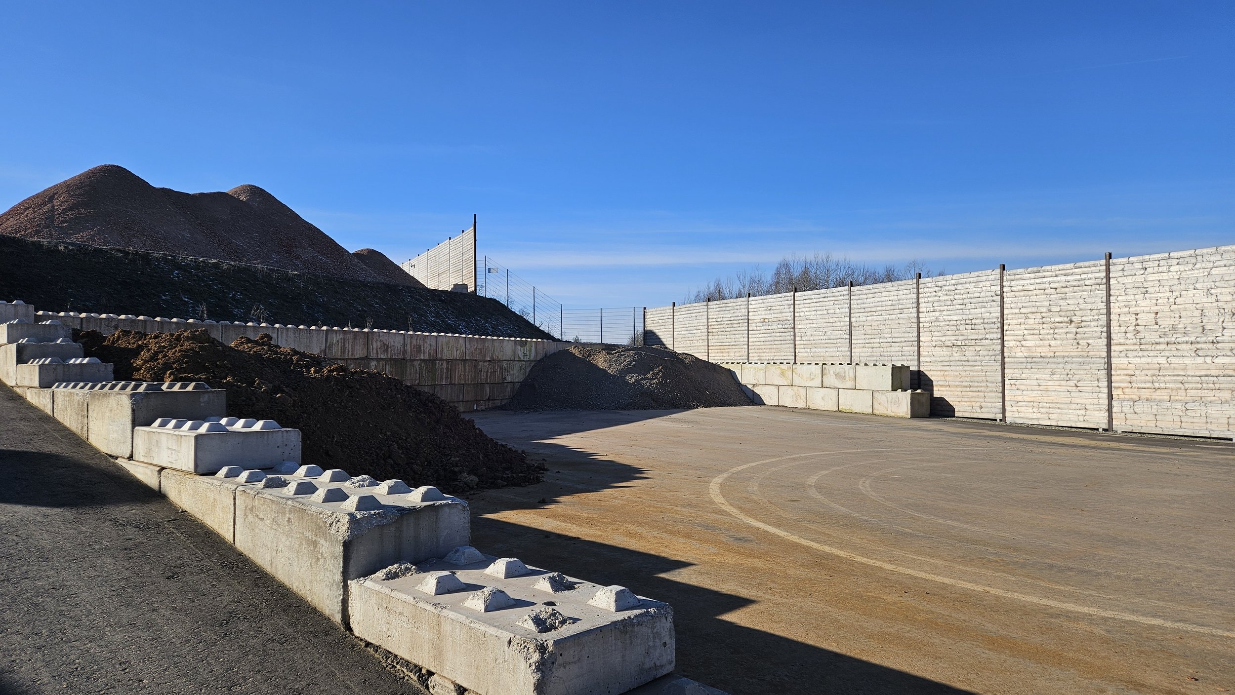 Ein sportlicher Fußballplatz in Bau mit Sand und Erde, umgeben von Bauzäunen und Steinmauern, Berge im Hintergrund und ein blauer Himmel.