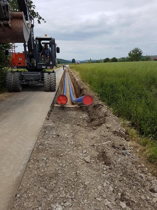 Bauarbeiter legt eine Wasserleitung an einer Landstraße neben einem Feld, mit einem großen Bagger im Hintergrund.