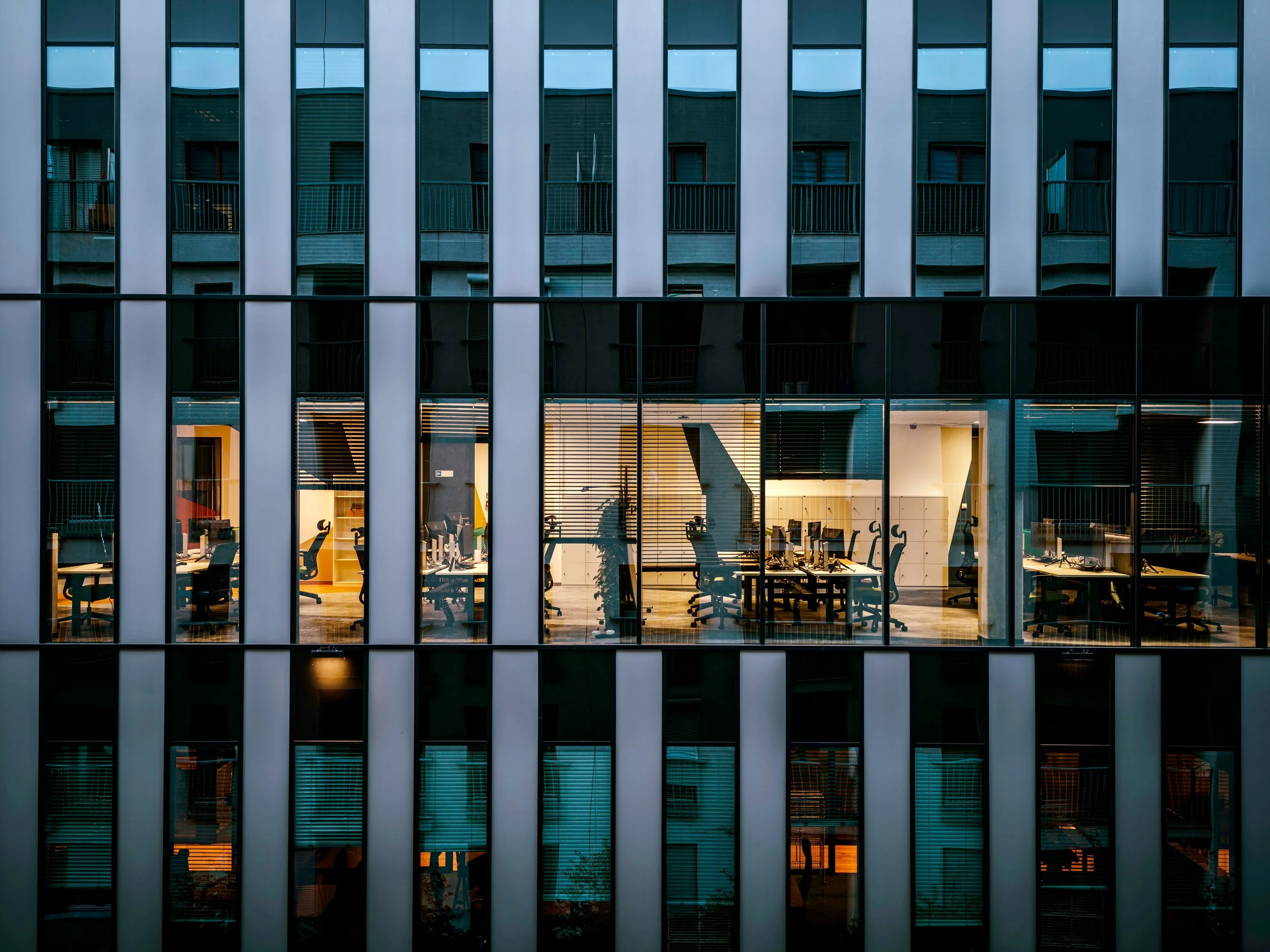Interior of an office building seen through large glass windows, with desks, chairs, and computers visible inside.