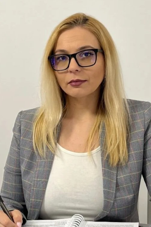 Andreea Iordache. A woman with blonde hair wearing glasses, a gray plaid blazer, and a white top, sitting at a desk with a notebook and pen, looking into the camera.