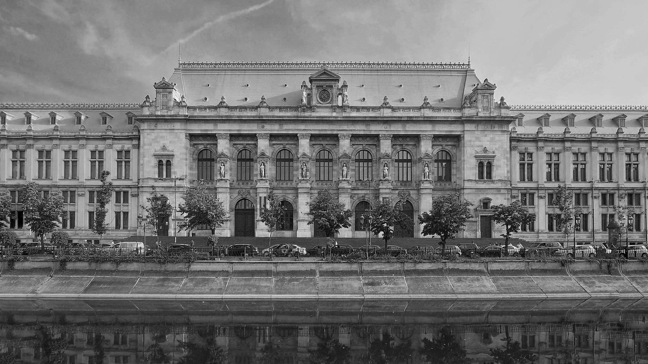 A historic building with classical architecture, large windows, and sculptures on the facade, situated behind a street with trees, parked cars, and a waterway reflecting the buildings.