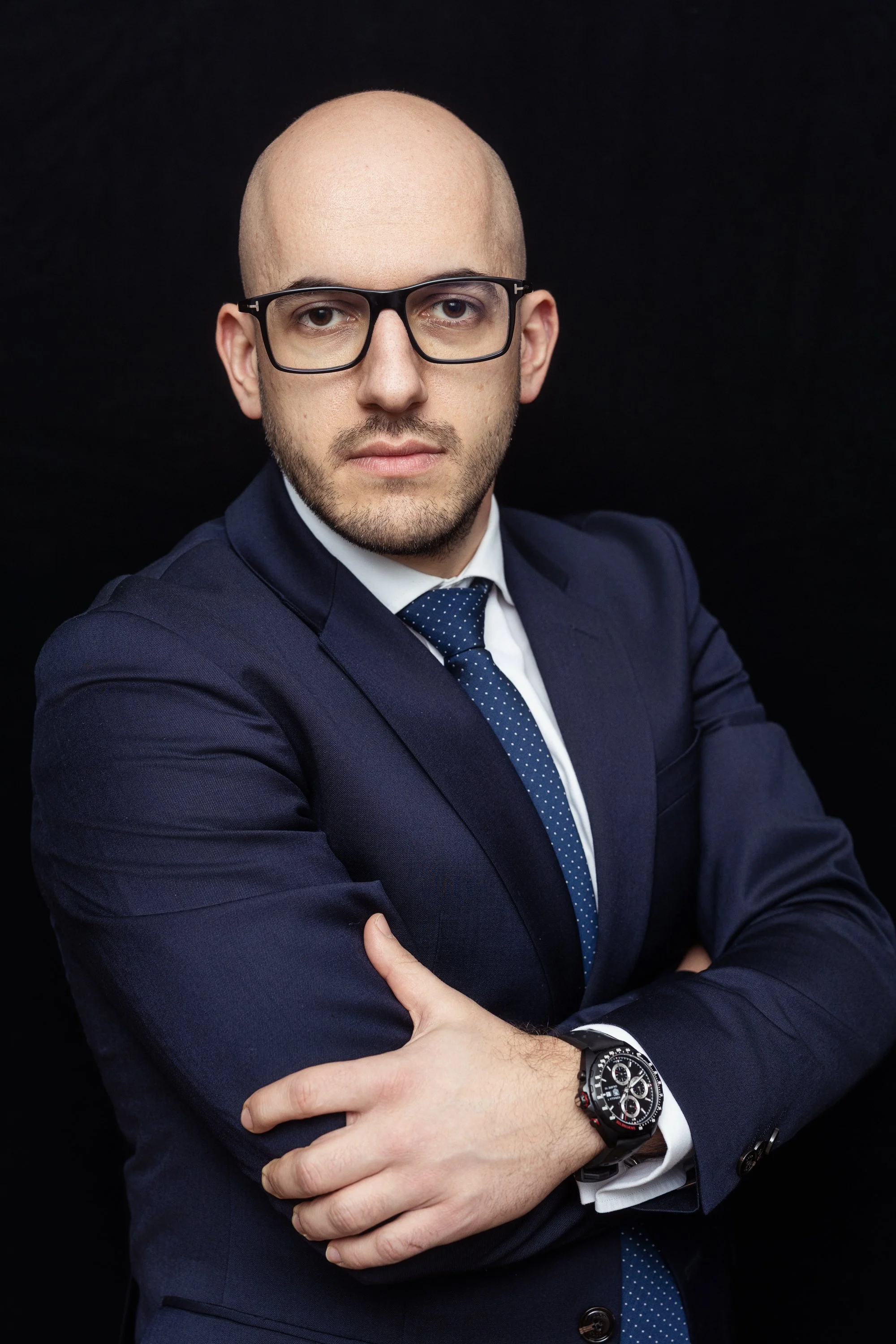 A professional man with glasses, dressed in a dark suit and tie, crossing his arms and looking confidently at the camera against a black background.