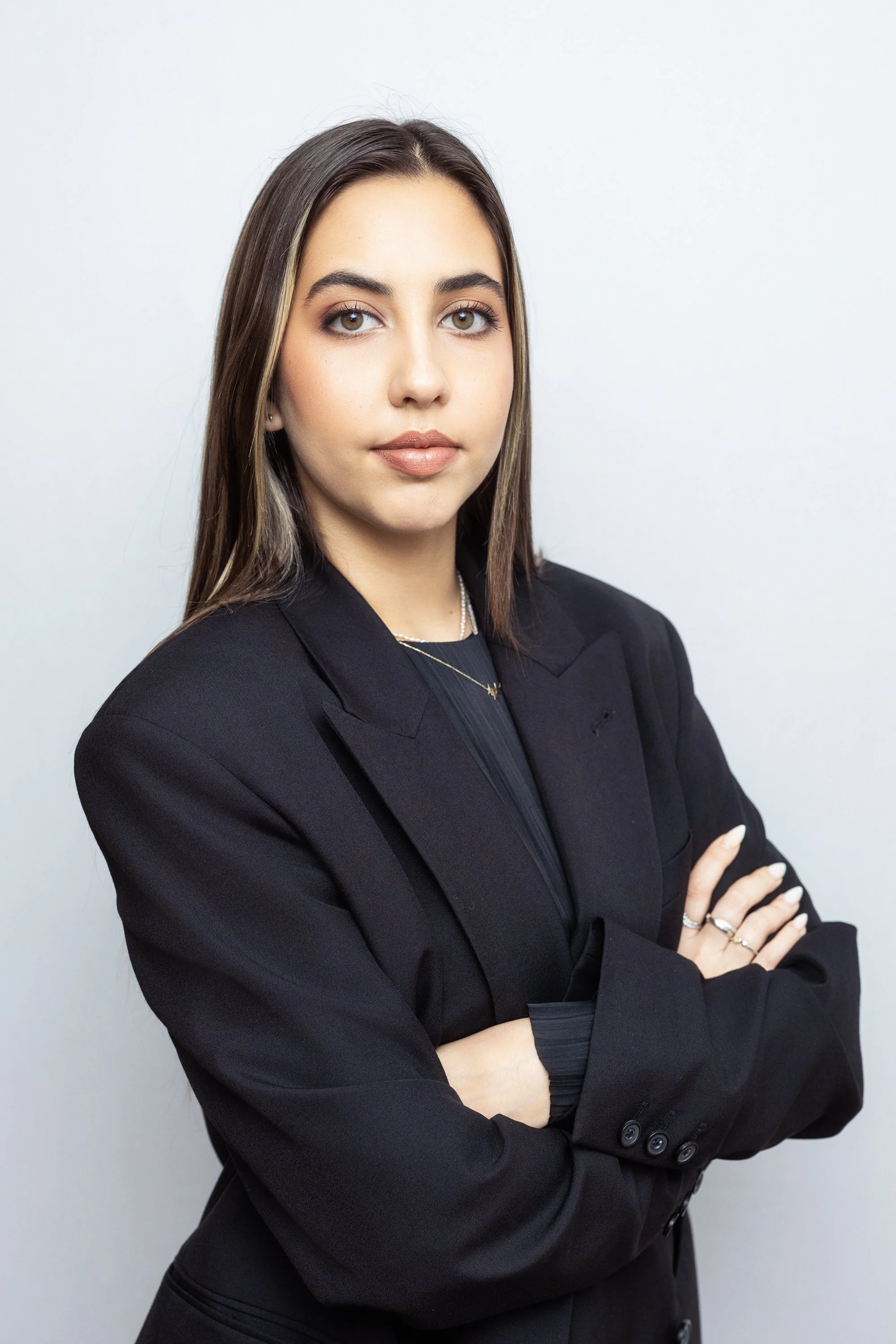 A woman with shoulder-length brown hair and light makeup, wearing a black blazer and jewelry, standing with her arms crossed against a plain light background.