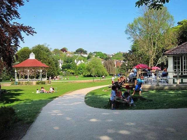 A park scene with a curved pathway, people relaxing on benches and grass, a white gazebo with a red roof, and a small building with red umbrellas, trees, and houses in the background on a sunny day.