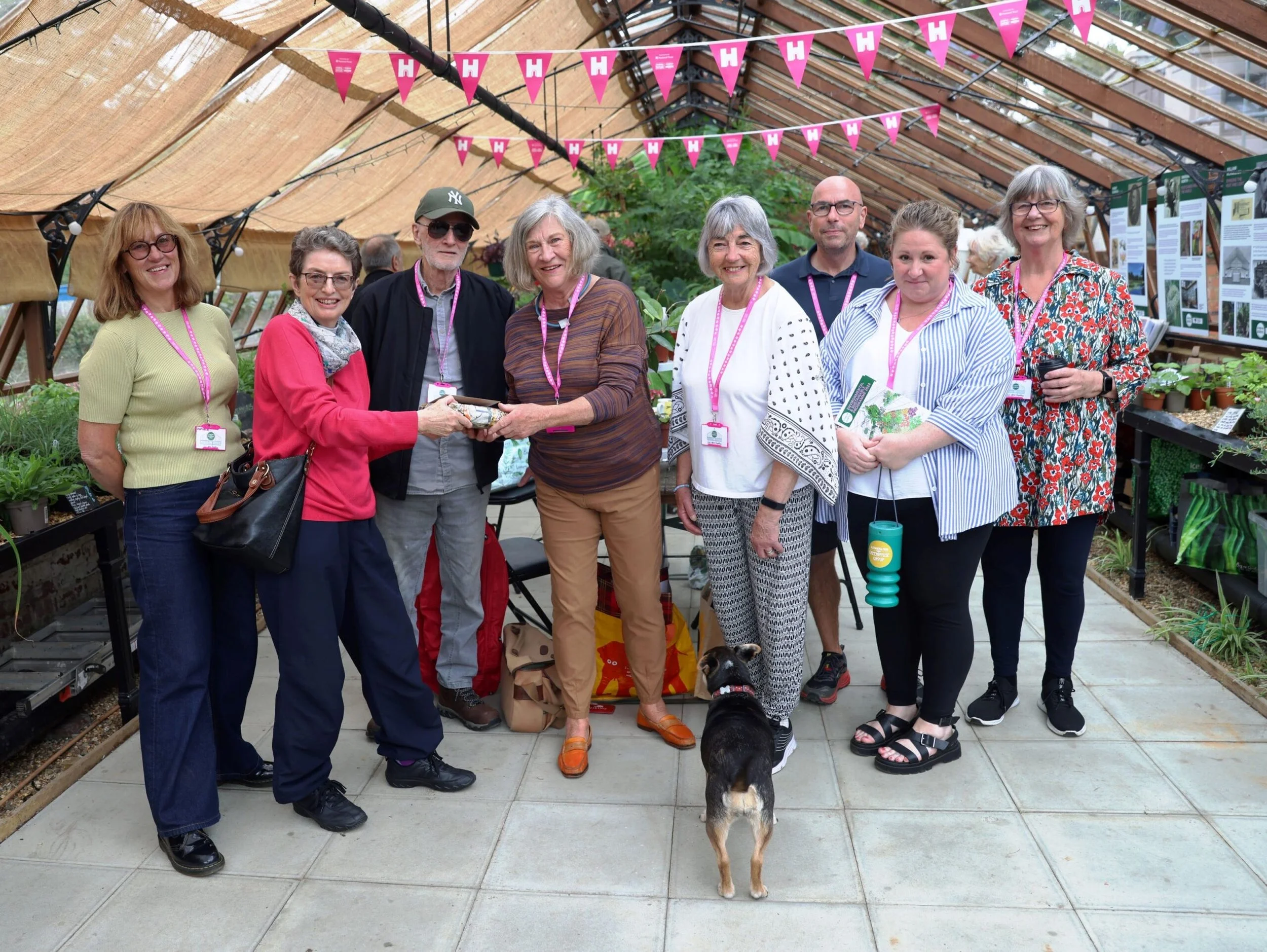 A group of nine people standing inside a greenhouse, with plants and informational posters around them, celebrating or participating in an event, with pink banners hanging overhead and a black dog at their feet.