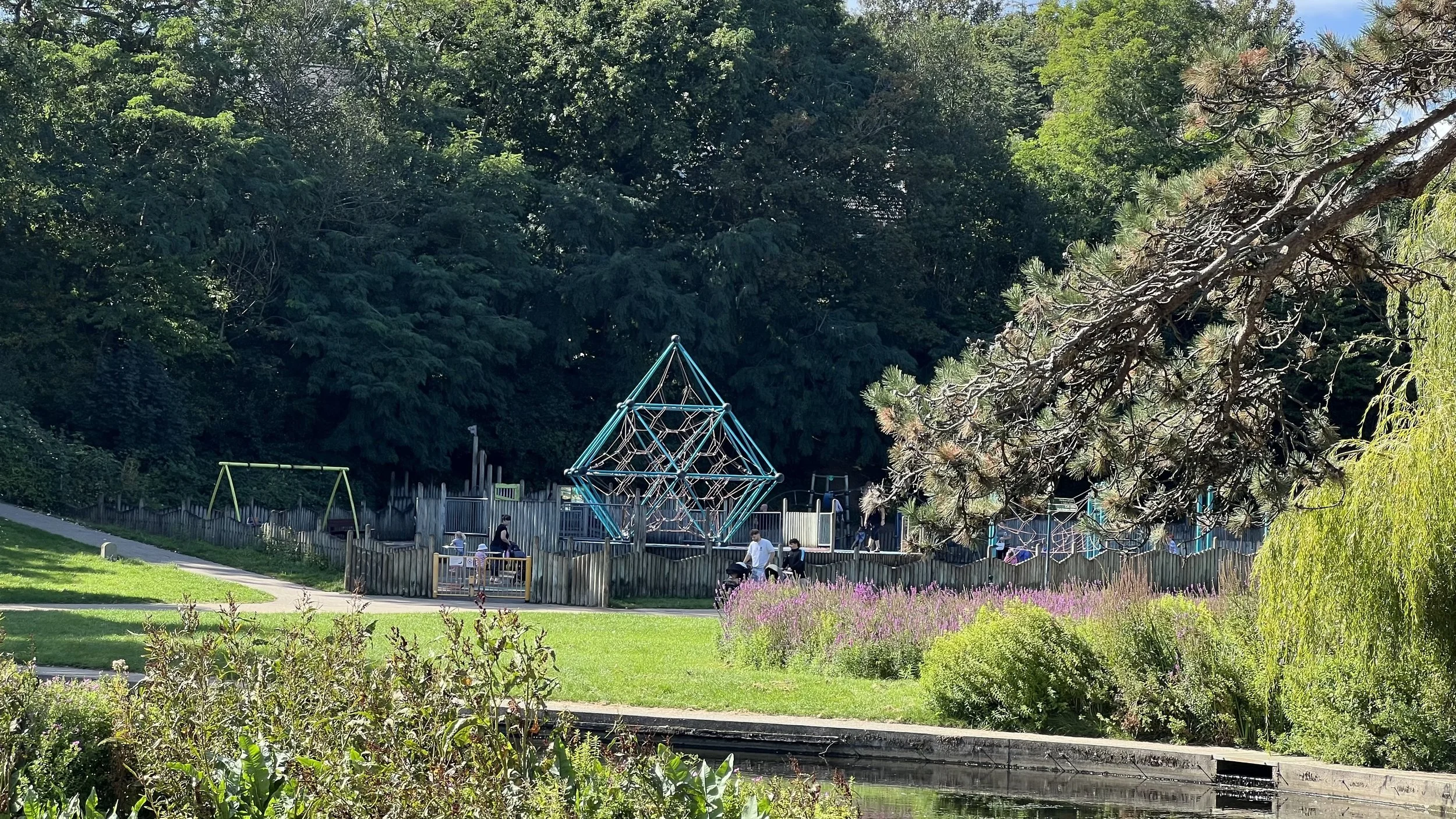 A park with a playground, a fence, and colorful flowers in the foreground, surrounded by tall green trees under a partly cloudy sky.