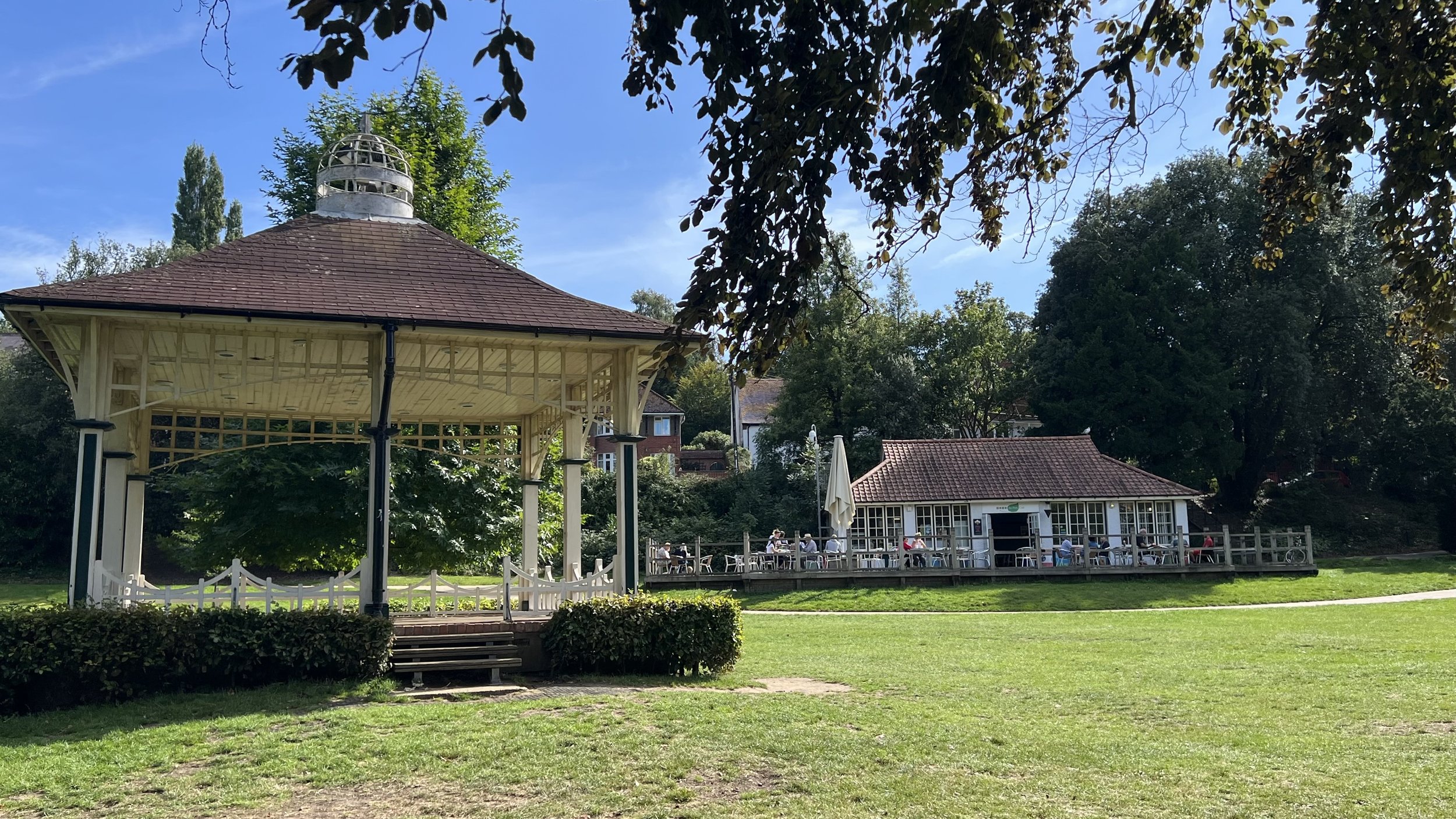A park scene with a large open grassy area, a gazebo structure with a red shingle roof, and a small building with outdoor seating where people are sitting at tables with umbrellas. There are trees around and a clear blue sky.