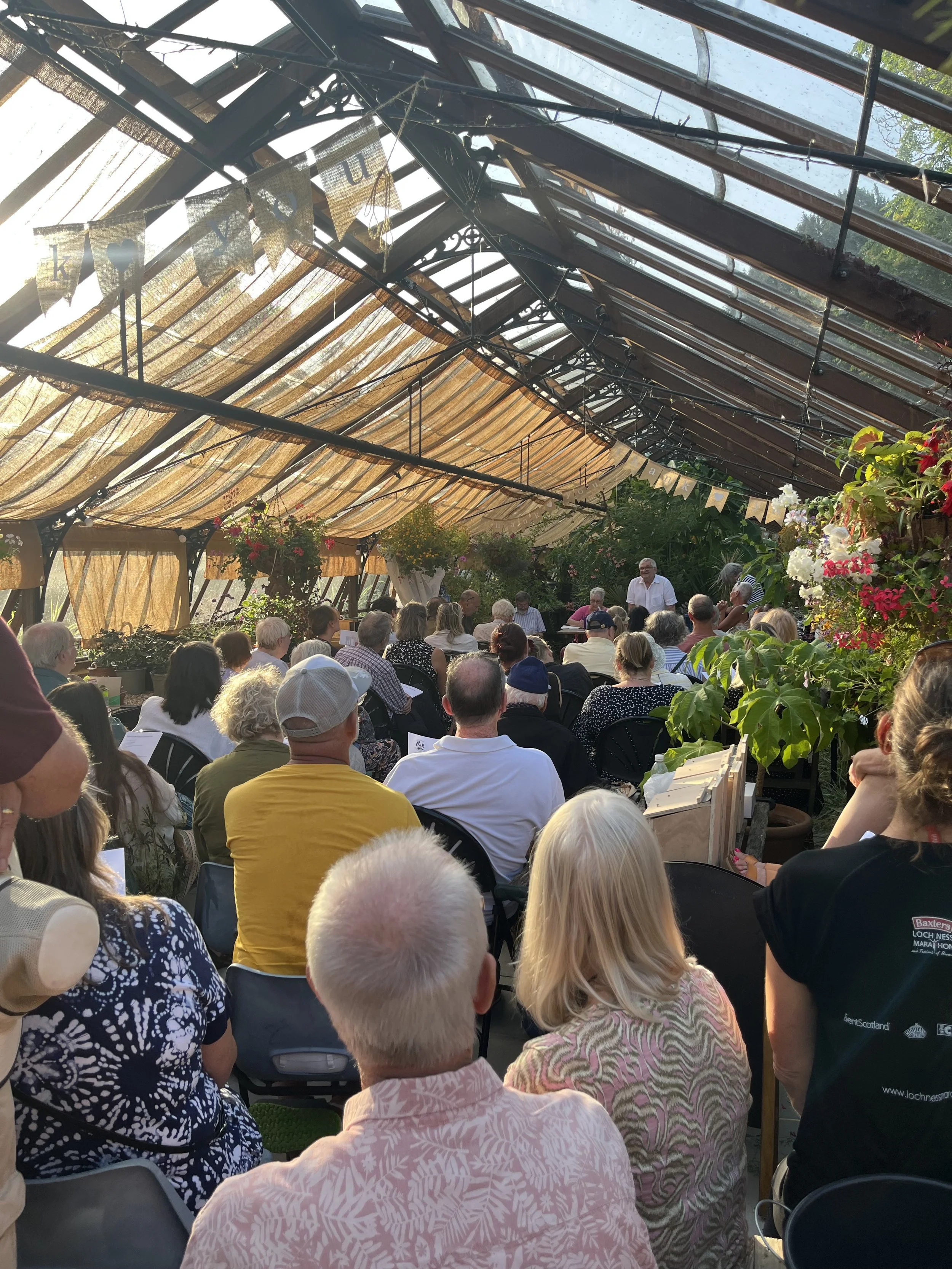 A large gathering of people seated under a glass greenhouse roof with colorful flowers and banners hanging, attending an outdoor event or presentation.