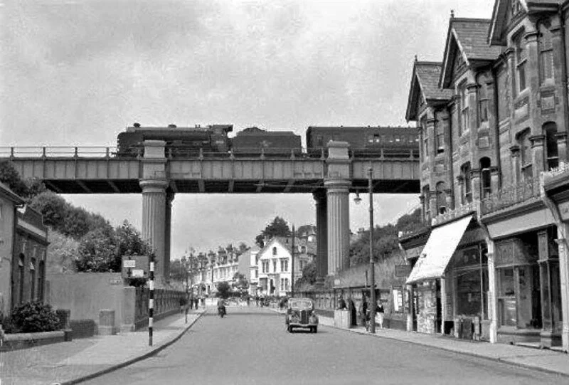 A black and white photo of a small town street with buildings on both sides, an elevated train track supported by large columns overhead, and a few vintage cars and pedestrians.