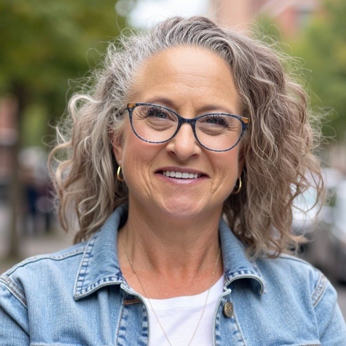A smiling woman with short, curly hair, wearing a black top and a striped jacket, standing in front of a glass wall.