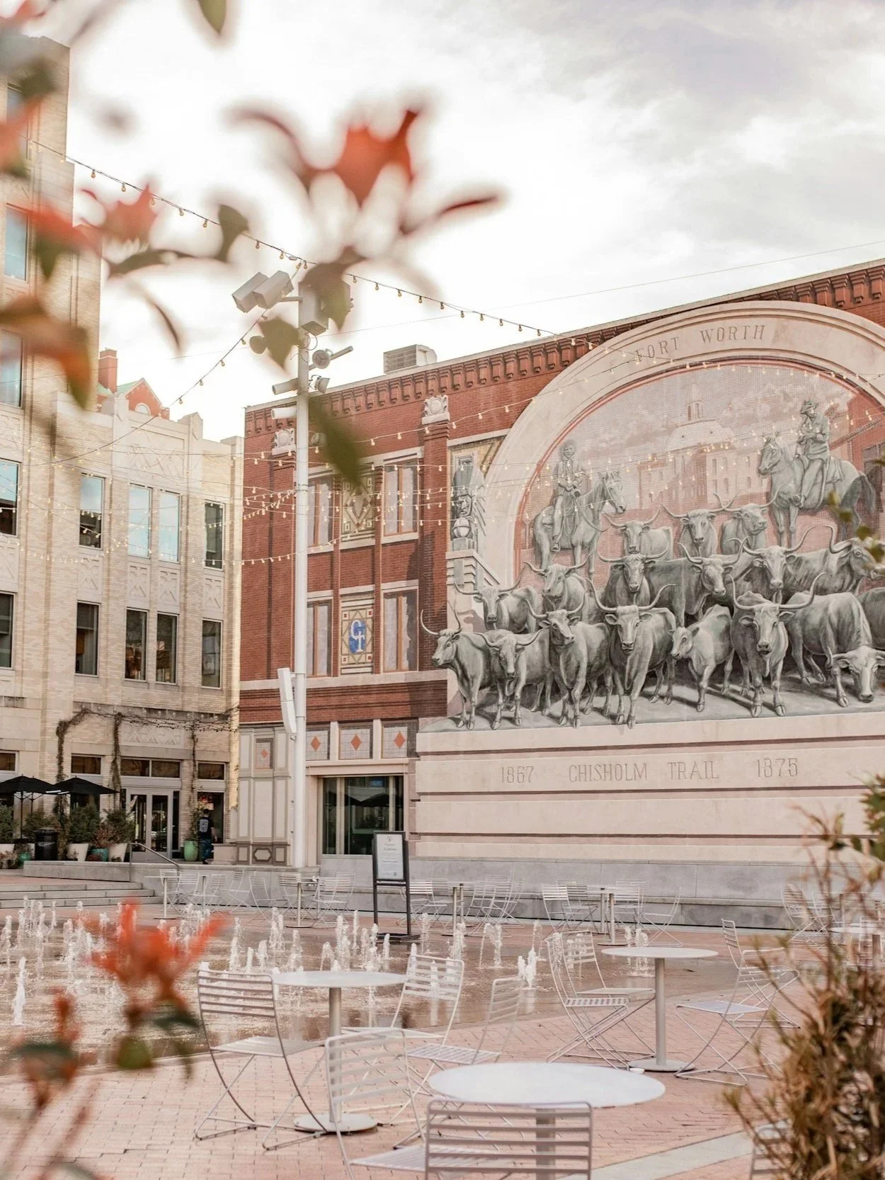 Sundance Square: An outdoor plaza with empty white tables and chairs, water fountain, historic mural on brick building, string lights, and potted plants, in Fort Worth, Texas.