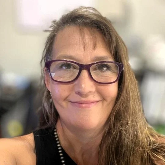 A woman with red hair, glasses, and a bright red blazer smiling in front of a dark, textured background.