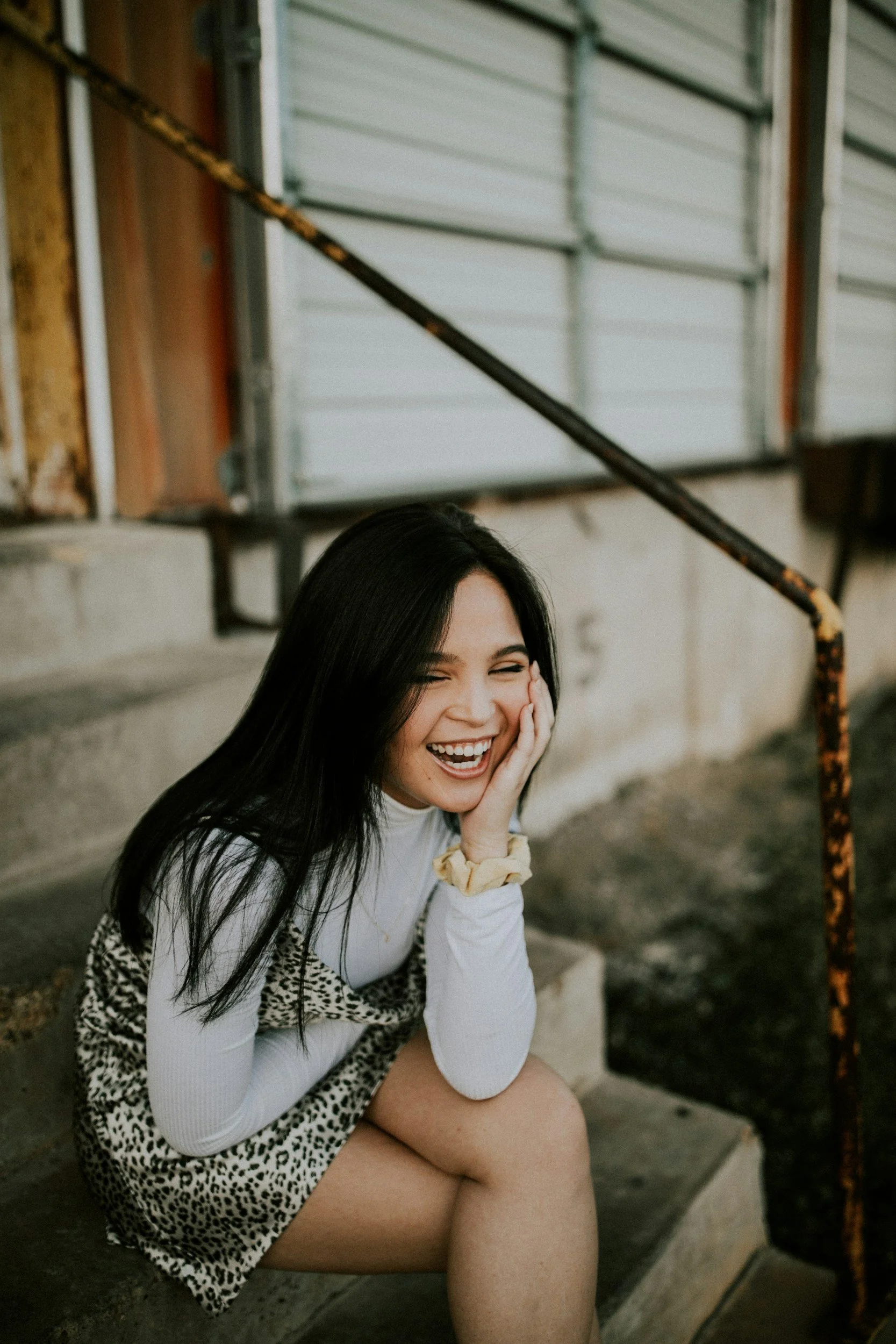 A young woman with long dark hair, wearing a white top and leopard print skirt, sitting on concrete steps outside, smiling and laughing.