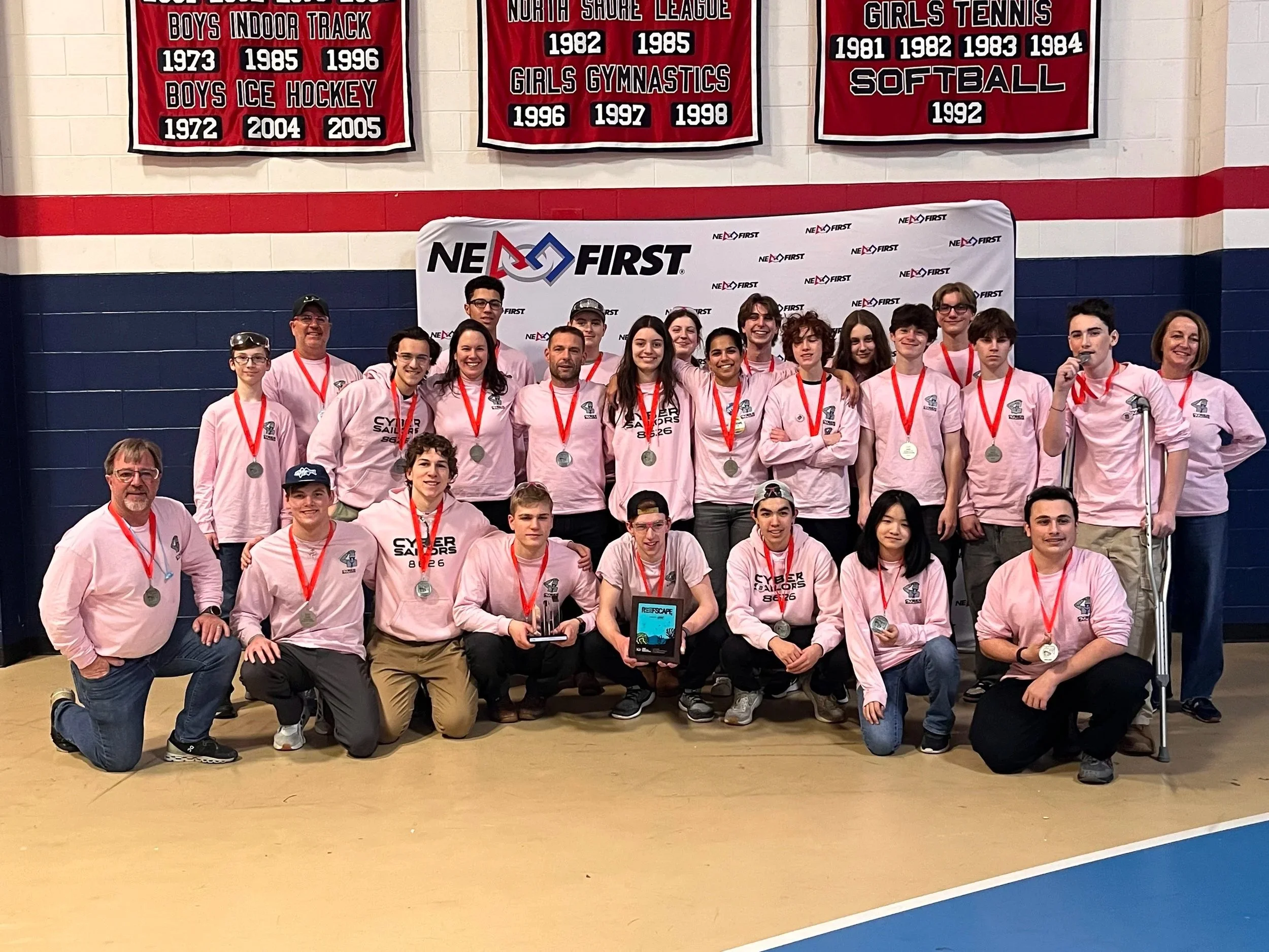 A group of students and coaches wearing pink shirts with medals around their necks posing in front of a NE FIRST banner at a robotics competition. The background has banner awards for various sports and activities.