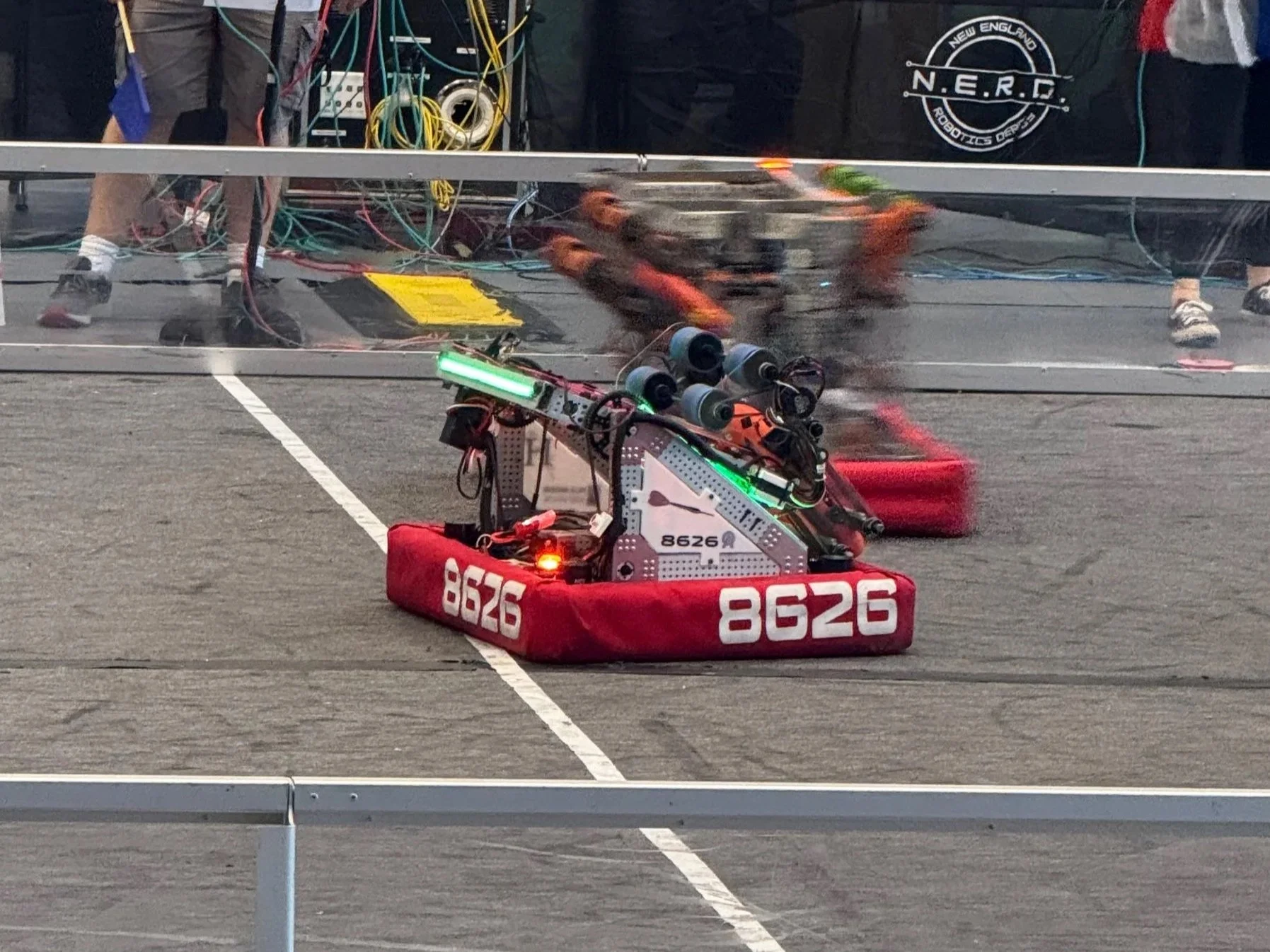 A robot with the number 8626 on its side is participating in a robotics competition, moving across a marked competition floor. The robot is built with various mechanical and electronic components, including a white structure, wiring, and sensors. In the background, a clear barrier separates the competition area from the spectators and team members.