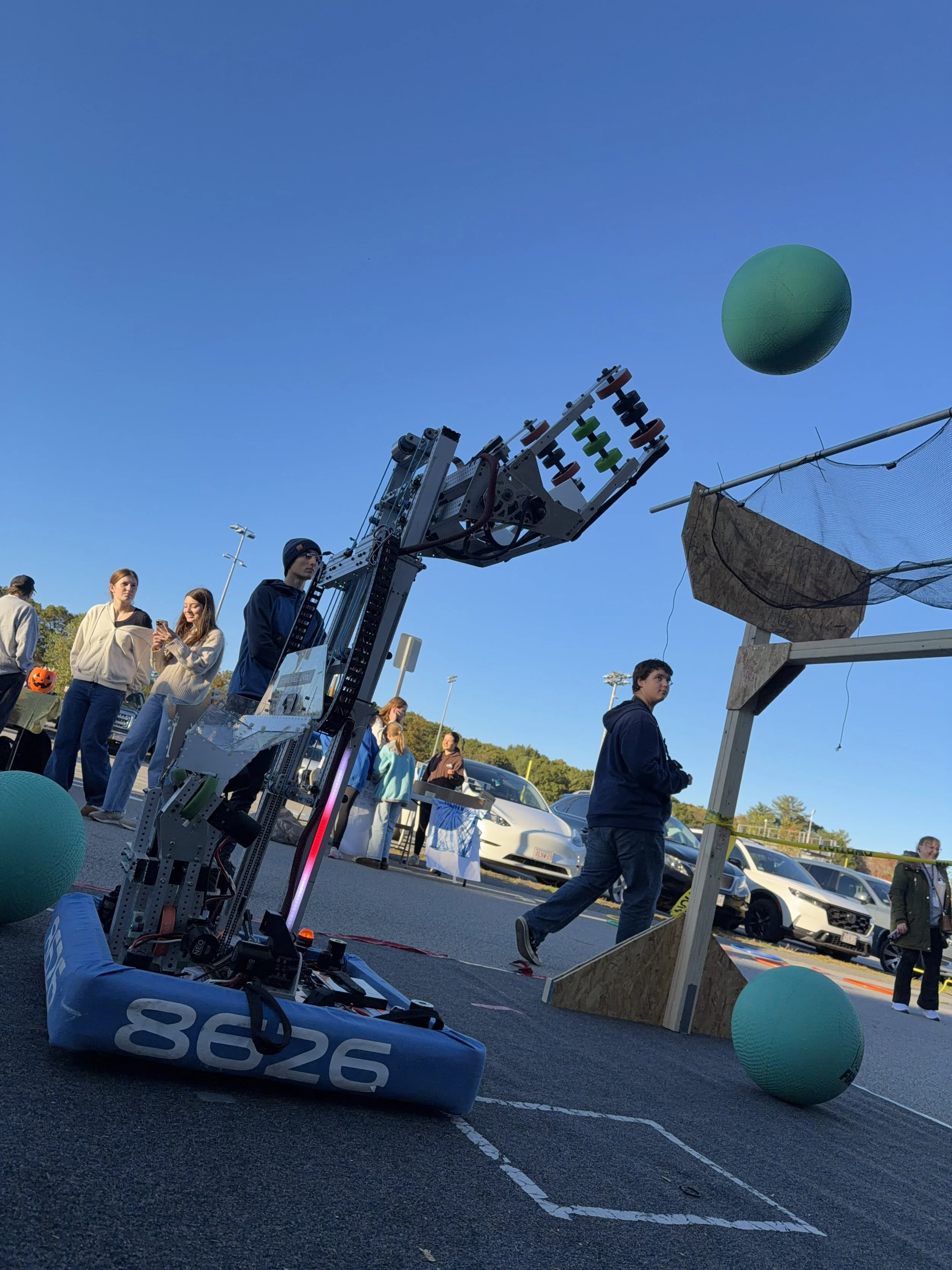 A robot is shooting a green ball into the air at an outdoor event with people watching and participating, parked cars, and clear blue sky in the background.