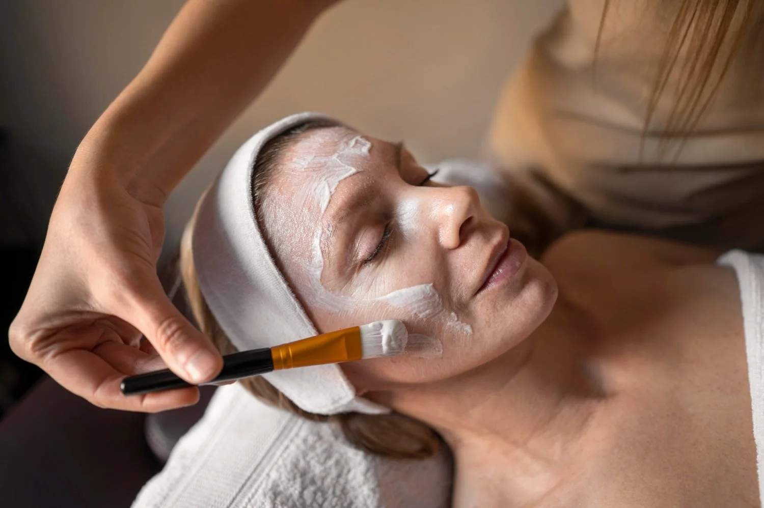 A woman receiving a facial treatment with white cream applied on her face, resting with eyes closed while a skincare professional uses a brush.
