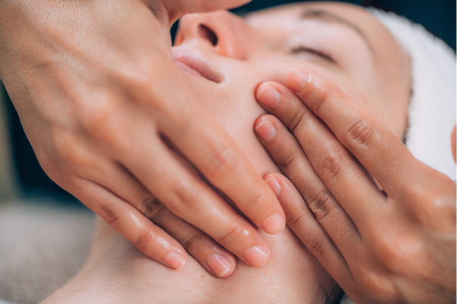 A woman receiving a facial treatment with white cream applied on her face, resting with eyes closed while a skincare professional uses a brush.