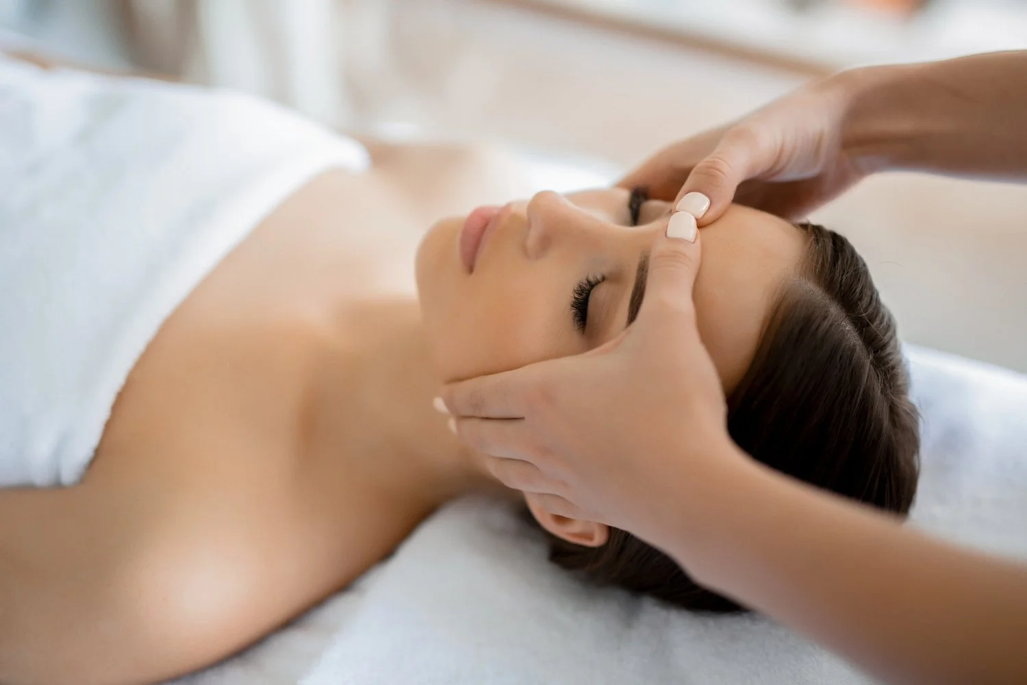 A woman receiving a facial massage in a spa, lying on a treatment table with her eyes closed.