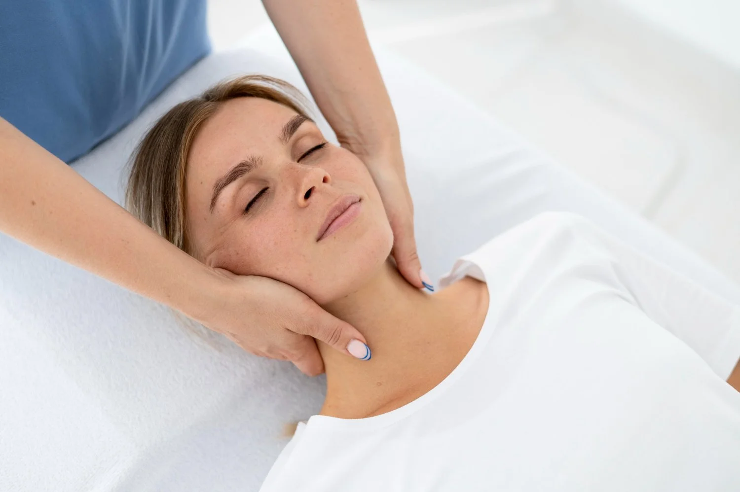 A woman relaxing with her eyes closed while someone gives her a neck massage. She appears comfortable on a white surface, possibly in a massage or therapy setting.