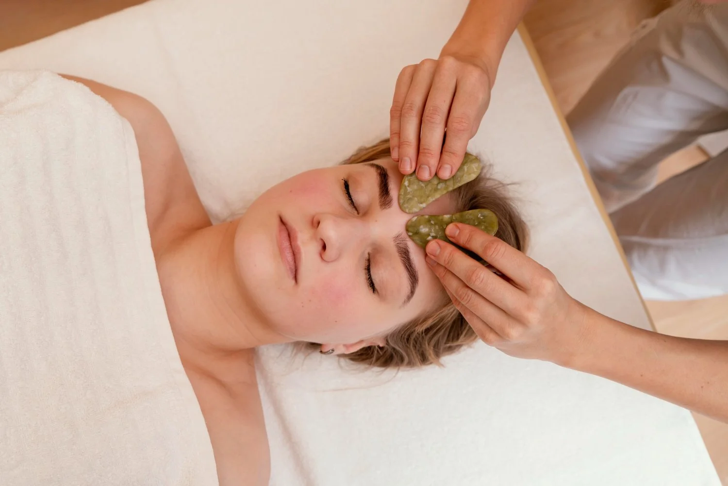 A woman receives a facial massage with jade stones on her forehead while lying on a treatment bed, eyes closed.