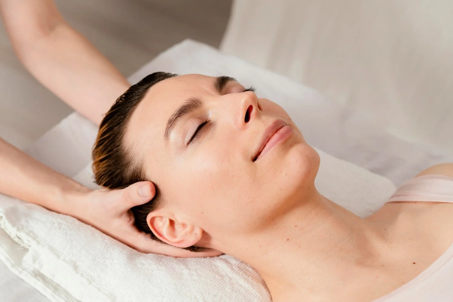 A woman receiving a facial massage while lying on a treatment bed with her eyes closed.