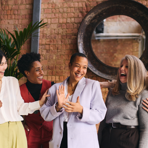 Four women laughing and smiling together indoors, with a brick wall and a large round mirror in the background.