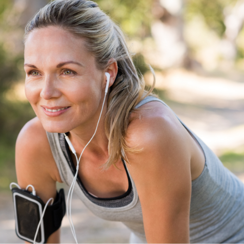 A woman with blonde hair smiling, wearing a gray athletic tank top and earbuds, outdoors in a park.
